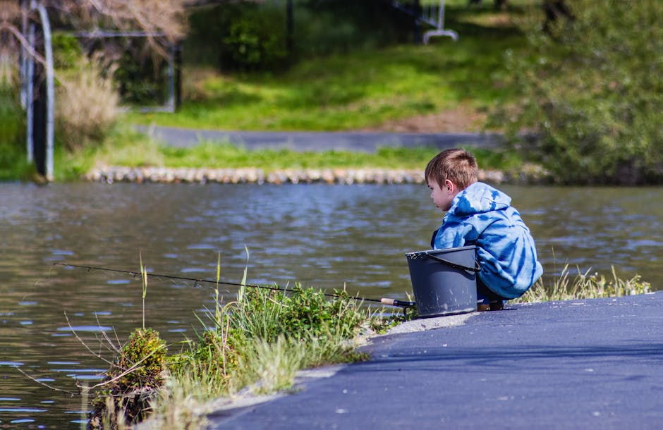 A young boy sitting by a lake, engaged in fishing on a sunny day in a park.